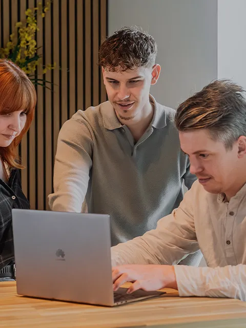 Drei Kollegen schauen gemeinsam auf einen Laptop; einer von ihnen zeigt auf etwas.